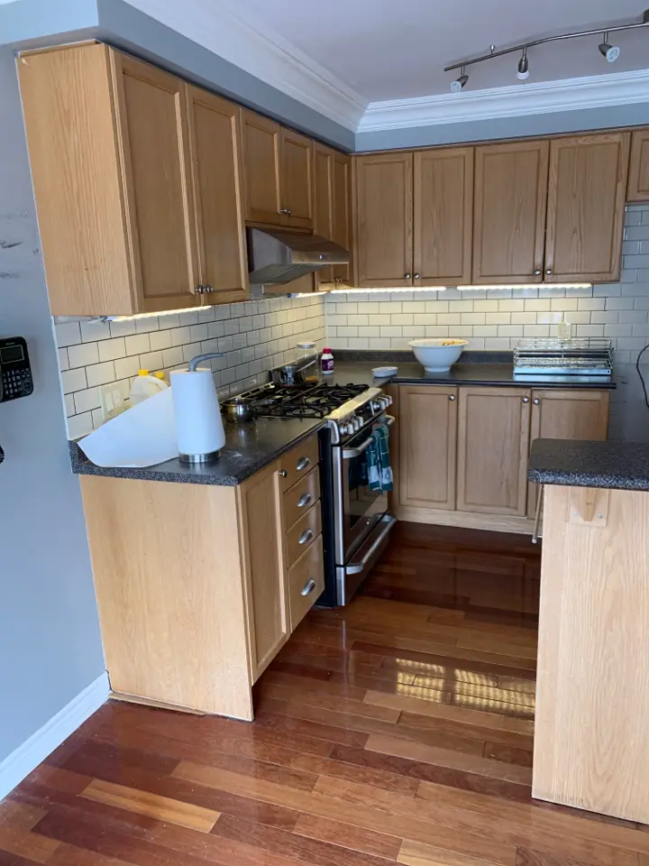 A before renovation kitchen with wooden cabinets, a tile backsplash, and a gas stove with stainless steel oven. There are bowls on the countertop and a roll of paper towels is placed in a holder. The flooring is wooden, and there's track lighting on the ceiling.