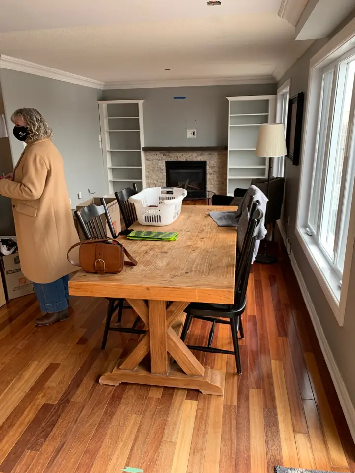 A wooden dining table in a before renovation living room with hardwood floors. A fireplace with white shelves on either side is in the background. The room has a large window on the right.