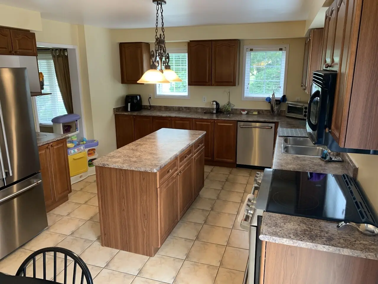 A before renovation kitchen with wooden cabinets, tile flooring, an island counter, stainless steel appliances including a refrigerator and a dishwasher, and two windows providing natural light.