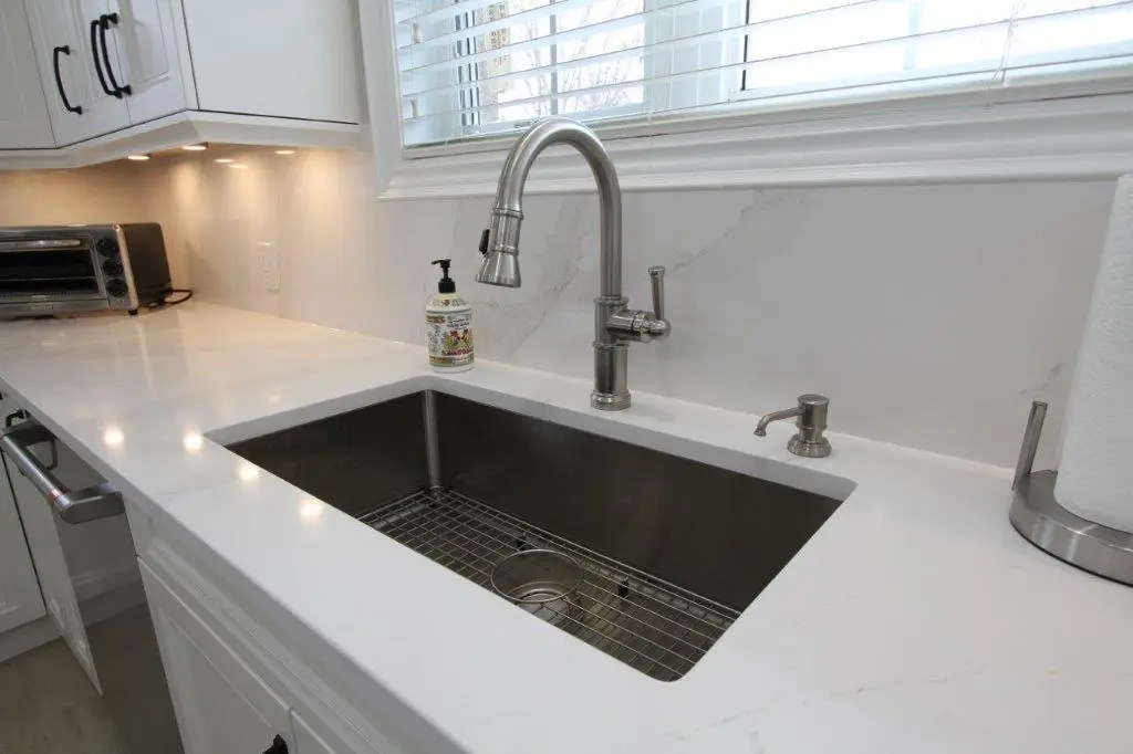 Modern kitchen sink with a stainless steel faucet, quartz countertop, and a soap dispenser nearby, under a window with blinds.