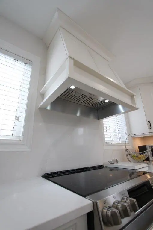 A modern kitchen with a white range hood above a stainless steel stove, flanked by two narrow windows with blinds.