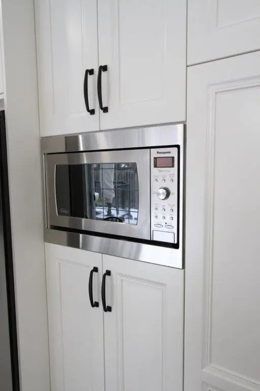 A stainless steel microwave oven installed within white kitchen cabinetry, featuring black handles on the surrounding cabinets.