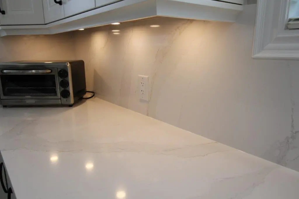 A modern quartz kitchen countertop with a polished, light-colored surface and a matching backsplash, featuring under-cabinet lighting and a compact stainless steel toaster oven.