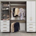 A neatly organized closet featuring hanging clothes including dresses and jackets, glass shelves displaying handbags and hats, and drawers with gold handles beneath.