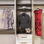 A wardrobe display featuring a floral dress, a black patterned jacket with a cap above, and a red and black floral dress. Beneath the jacket are a black quilted handbag and a pair of black high heels.