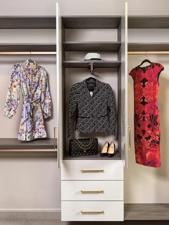 A wardrobe display featuring a floral dress, a black patterned jacket with a cap above, and a red and black floral dress. Beneath the jacket are a black quilted handbag and a pair of black high heels.