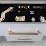 An open drawer containing neatly organized jewelry and accessories, including a pearl necklace, a diamond ring, designer brooch, watch, and sunglasses.