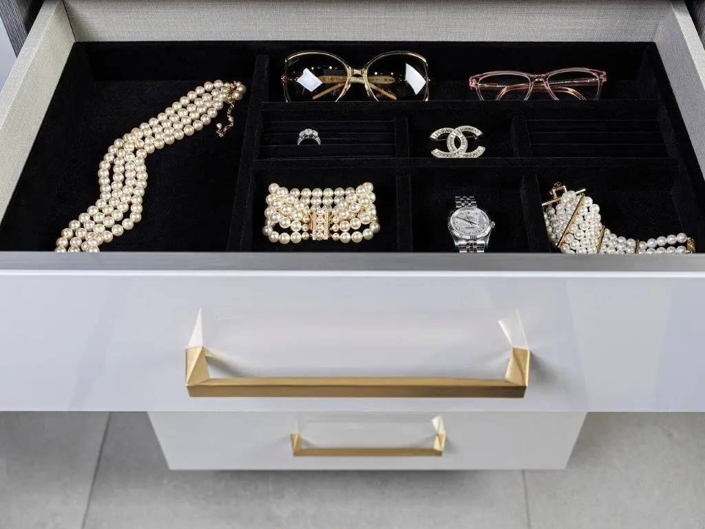 An open drawer containing neatly organized jewelry and accessories, including a pearl necklace, a diamond ring, designer brooch, watch, and sunglasses.