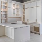 A modern home office space featuring white cabinetry with sleek handles, built-in shelves displaying decorative items, and a white desk with a chair. Overhead, track lighting is installed on the ceiling, illuminating the workspace.