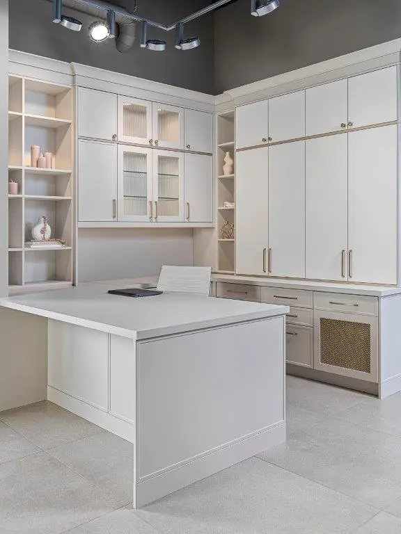 A modern home office space featuring white cabinetry with sleek handles, built-in shelves displaying decorative items, and a white desk with a chair. Overhead, track lighting is installed on the ceiling, illuminating the workspace.