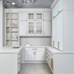 A modern home office with white cabinetry featuring glass-front upper cabinets, open shelves with decorative items, sleek countertops, and brushed metal handles. The ceiling has exposed ductwork and track lighting.