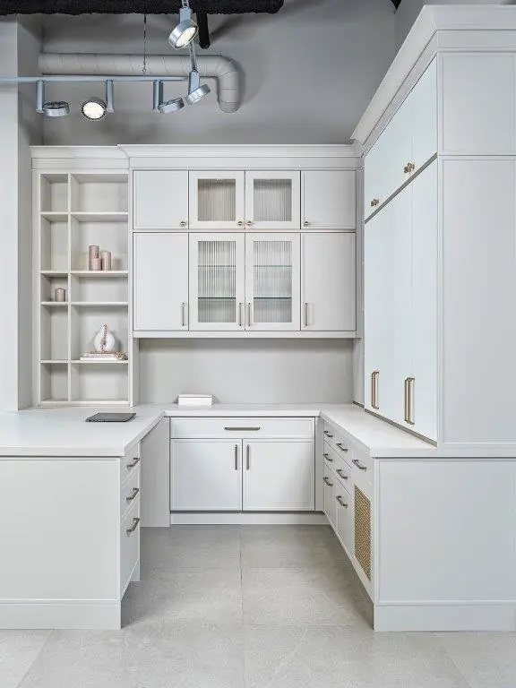A modern home office with white cabinetry featuring glass-front upper cabinets, open shelves with decorative items, sleek countertops, and brushed metal handles. The ceiling has exposed ductwork and track lighting.