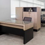 A modern office space featuring two wooden desks with black accents and glass tops, accompanied by mesh office chairs and matching storage cabinets against a windowed wall.