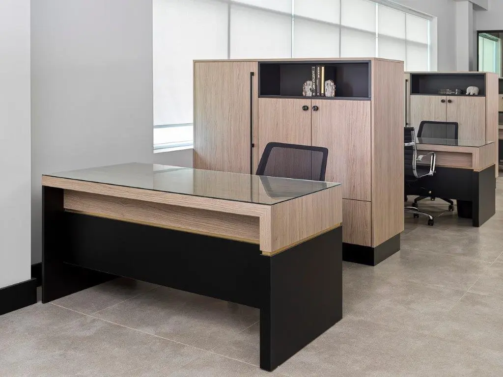 A modern office space featuring two wooden desks with black accents and glass tops, accompanied by mesh office chairs and matching storage cabinets against a windowed wall.