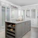 A luxurious walk-in closet with a central gray storage island featuring a marble countertop and shoe shelves, surrounded by white cabinetry with glass doors and neatly organized clothes.