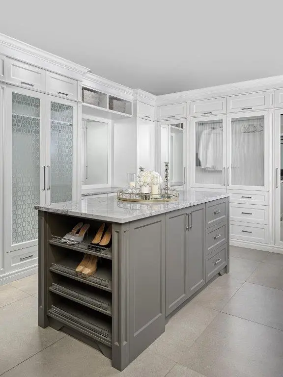A luxurious walk-in closet with a central gray storage island featuring a marble countertop and shoe shelves, surrounded by white cabinetry with glass doors and neatly organized clothes.