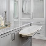 A modern closet with grey cabinetry featuring a pull-out ironing board and a marble-topped island with decorative items on a tray.
