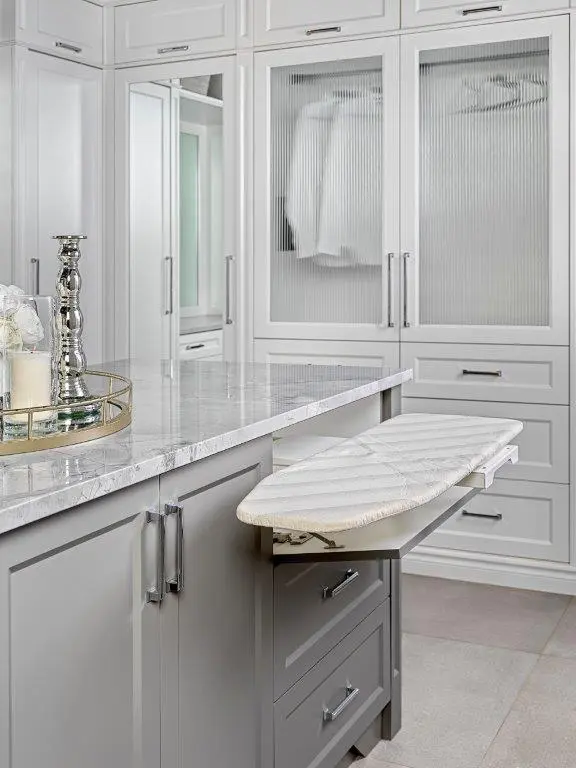 A modern closet with grey cabinetry featuring a pull-out ironing board and a marble-topped island with decorative items on a tray.