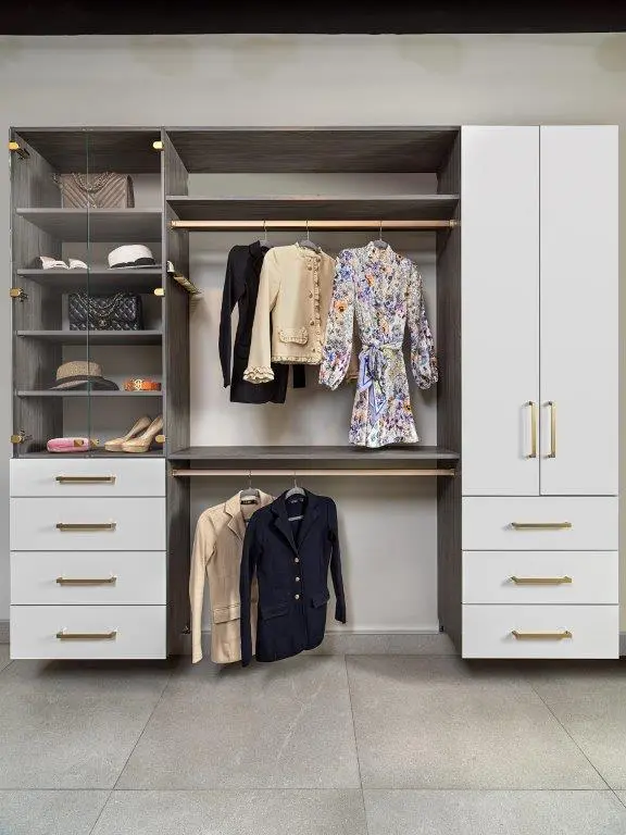 A modern closet featuring white and wood shelving with hanging rods, displaying jackets and a floral dress, alongside shelves with handbags, hats, and shoes.