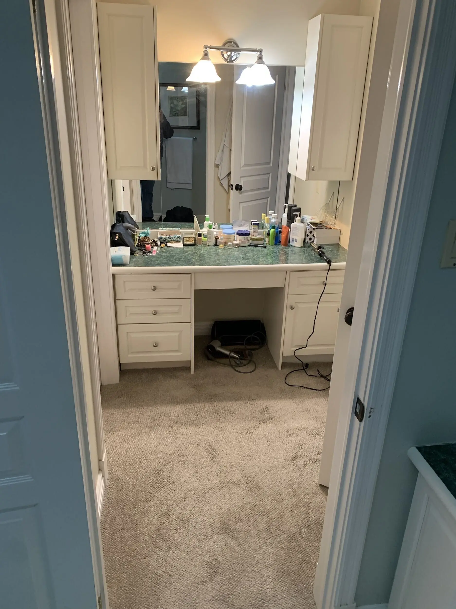 A before renovation bathroom vanity area with a green countertop. There are two wall-mounted cabinets, a large mirror, and overhead lights illuminating the space.