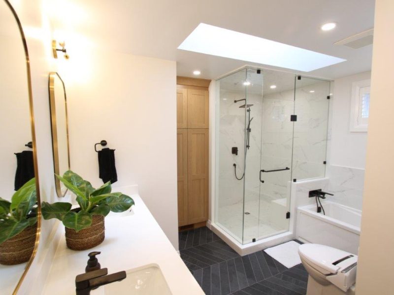 Modern bathroom with a glass-enclosed shower, white porcelain walls, black fixtures, and a white vanity with a plant in a basket.