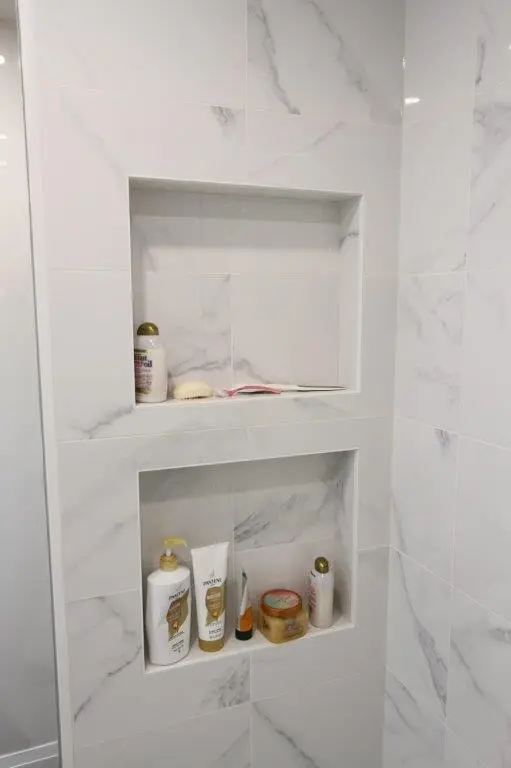 A shower wall with two recessed shelves, each holding shower products, against a backdrop of white porcelain-patterned tiles.