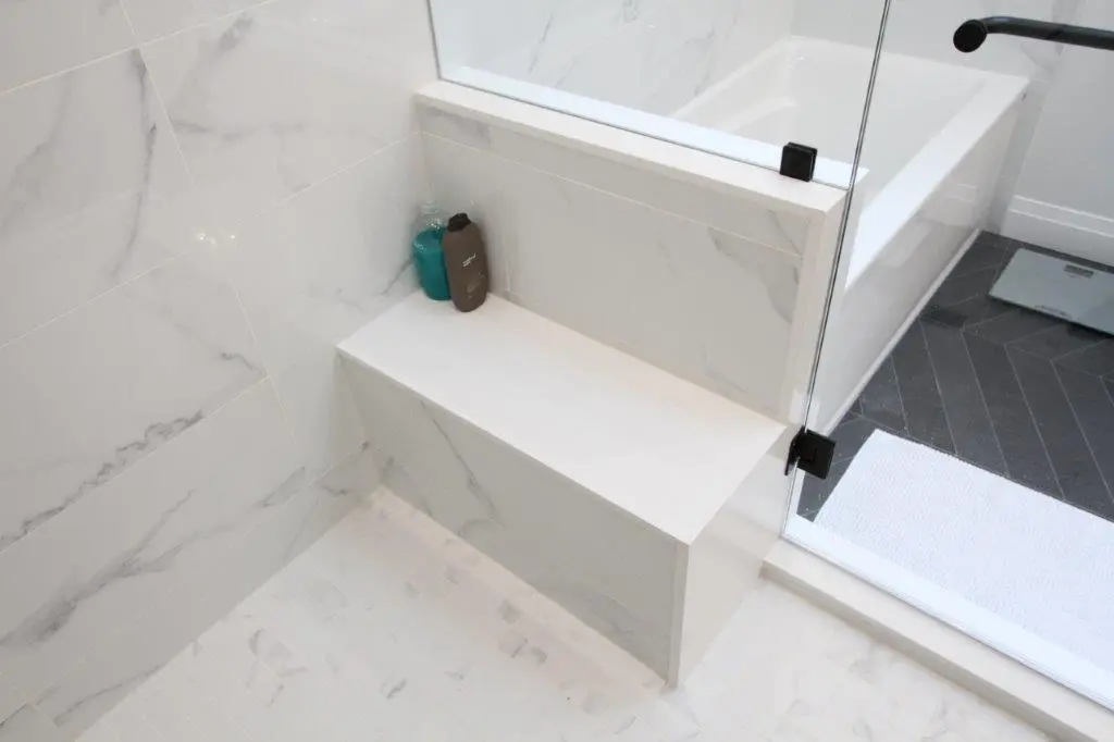 Modern bathroom featuring a glass shower enclosure with marble-patterned walls and a built-in shower bench and dark tile flooring is partially visible outside the enclosure.