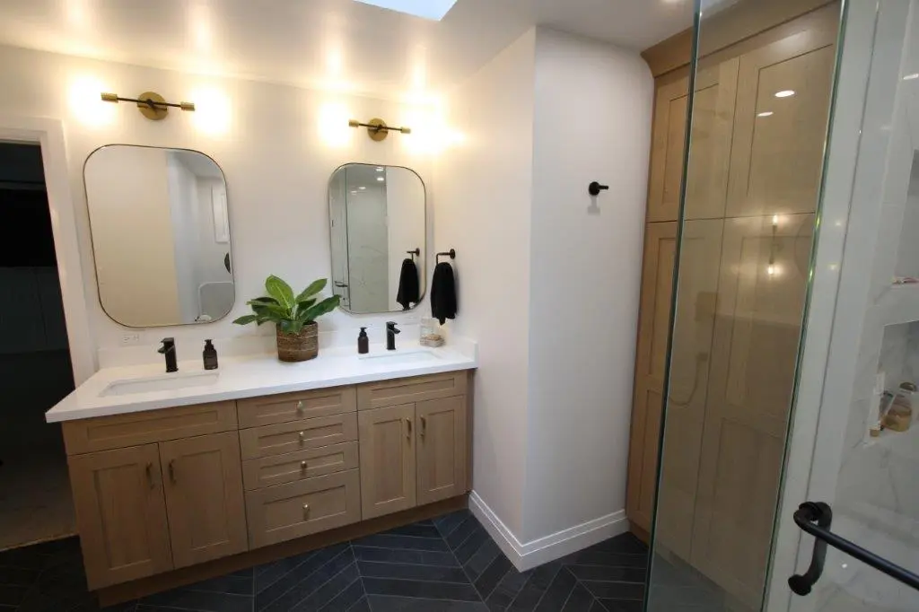 Modern bathroom with a double vanity featuring wooden cabinets, two mirrors with overhead lighting, and a potted plant as decoration. Adjacent is a glass-enclosed shower area.