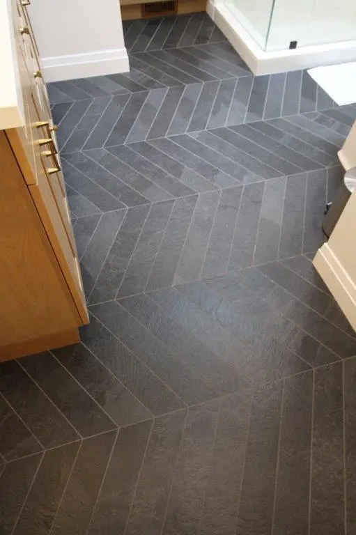 A bathroom featuring a dark herringbone patterned tile floor, with a wooden cabinet on the left and a glass shower enclosure on the right.