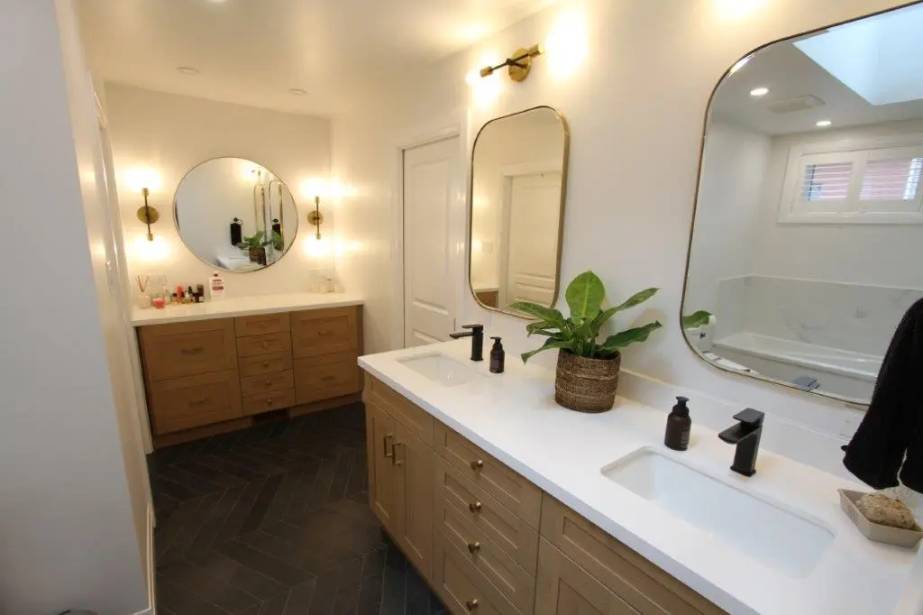 A modern bathroom with two wooden vanities featuring white quartz countertops and black faucets. Each vanity is equipped with a mirror, and the room is well-lit with wall sconces. A potted plant decorates one of the countertops, and a window provides natural light.