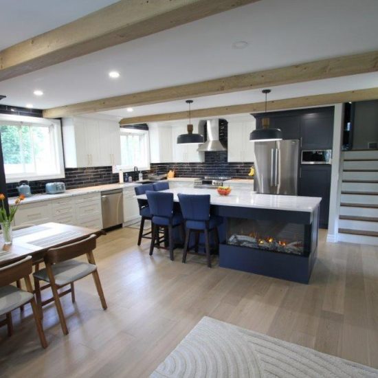 A modern kitchen with light wood flooring, featuring a central island with a built-in fireplace and blue chairs. The kitchen has black and white cabinetry, stainless steel appliances, and is accented by wooden ceiling beams. A dining table with chairs and a vase of flowers is located in the left foreground, and a staircase is visible in the background.