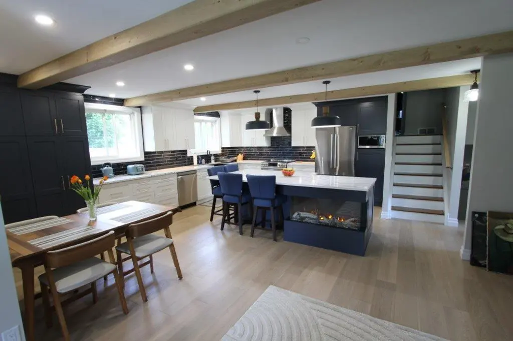 A modern kitchen with light wood flooring, featuring a central island with a built-in fireplace and blue chairs. The kitchen has black and white cabinetry, stainless steel appliances, and is accented by wooden ceiling beams. A dining table with chairs and a vase of flowers is located in the left foreground, and a staircase is visible in the background.