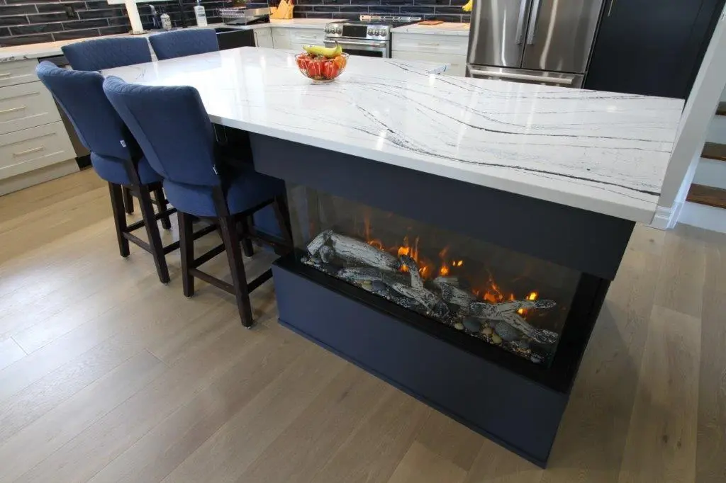 A modern kitchen island with a built-in electric fireplace, featuring dark blue chairs and a light-colored quartz countertop with subtle dark veining. The island is accompanied by a fruit bowl and is situated on light wood flooring.