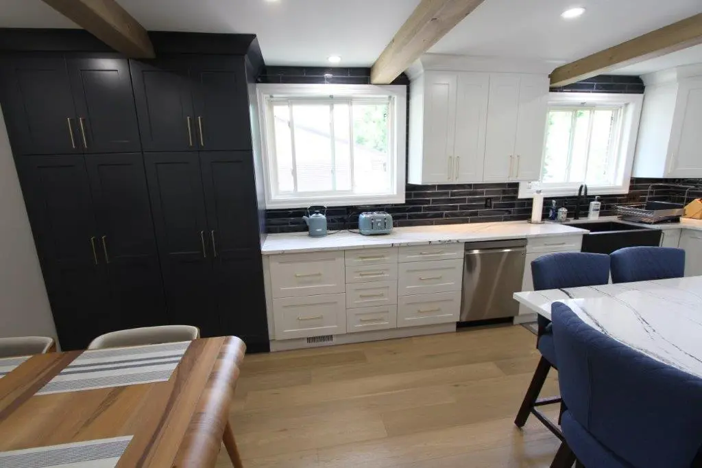 Modern kitchen with white cabinets, black backsplash, wood beams on the ceiling, and a wooden dining table. There are blue chairs, a stainless steel dishwasher, and a countertop with small kitchen appliances.