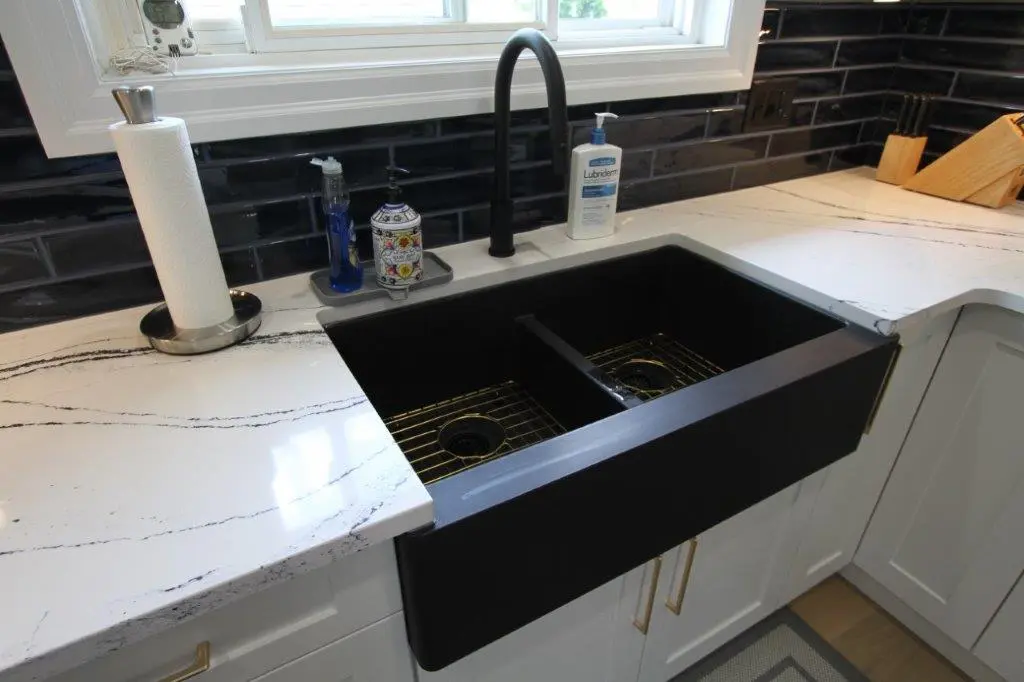 A modern kitchen with a black farmhouse sink and black faucet, set in a white countertop with gray veining. A soap dispenser and paper towel holder are on the counter, with black tiled backsplash and light filtering in from a window above.
