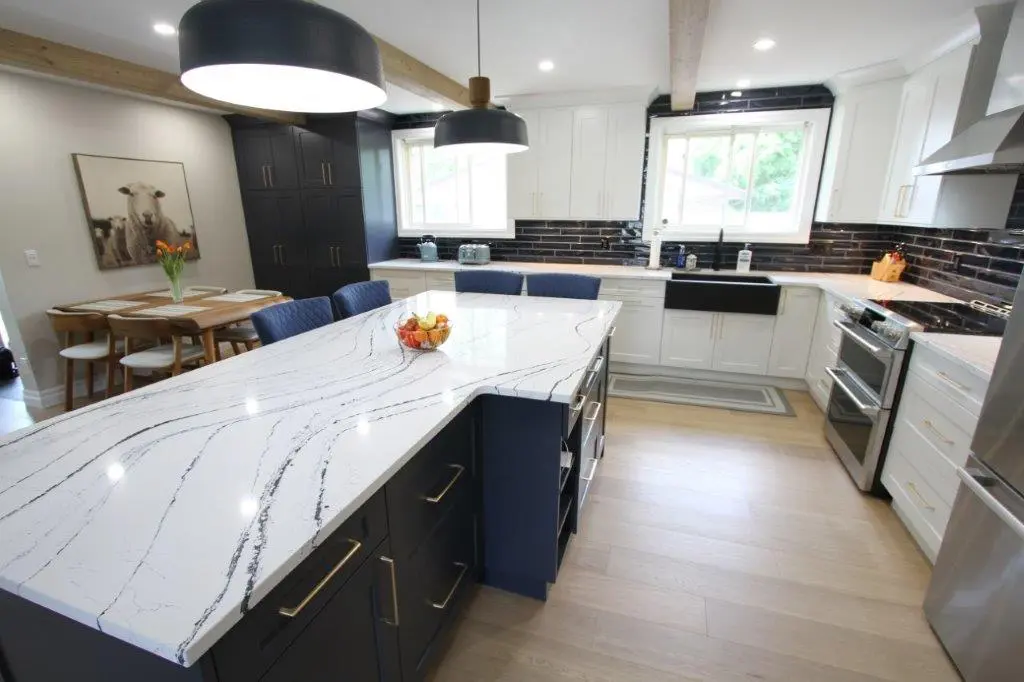 Modern kitchen with a large white marble island, dark cabinets, white cabinetry, stainless steel appliances, and a dining area with a wooden table and chairs. A large window provides natural light, and decorative elements include a bowl of fruit and a sheep-themed artwork on the wall.