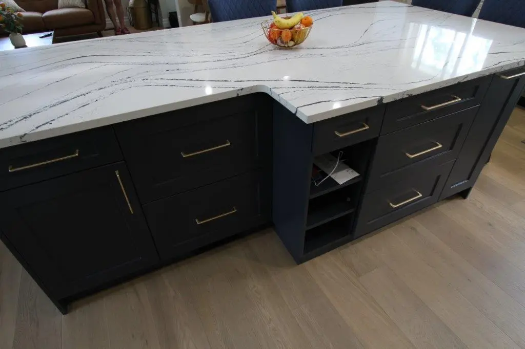 A kitchen island with a white quartz countertop featuring black veining, black cabinetry with gold handles, and an open shelving unit. A bowl of fruit sits atop the countertop.