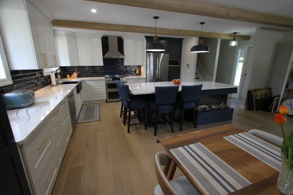 A modern kitchen with white cabinetry and black countertops, featuring a large kitchen island with three blue chairs. The kitchen has black tile backsplash, wooden flooring, and two pendant lights hanging above the island. A dining table with striped placemats is in the foreground, and there is a doorway to the outside in the background.
