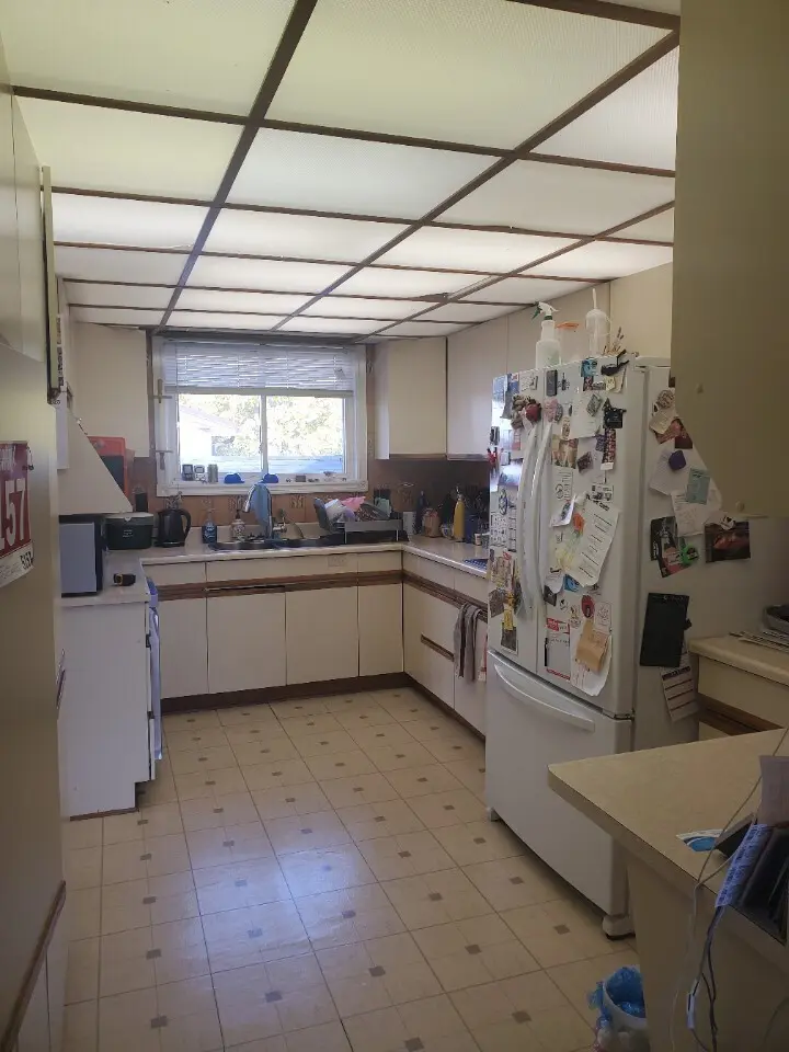 A before renovation kitchen with a tiled ceiling, brown and beige cabinets, a window above the sink, and a refrigerator covered with notes and magnets.