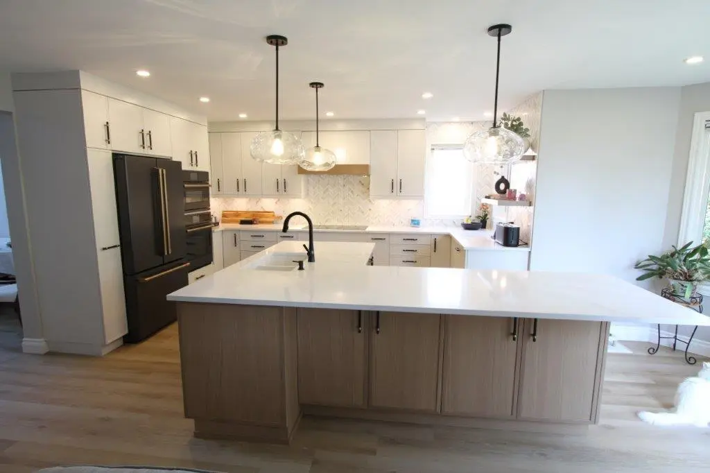 A modern kitchen with a large island featuring a sleek white quartz countertop, pendant lights hanging above, stainless steel appliances, and light wooden cabinetry. The room is bright, with plants near a window and a herringbone-patterned backsplash.