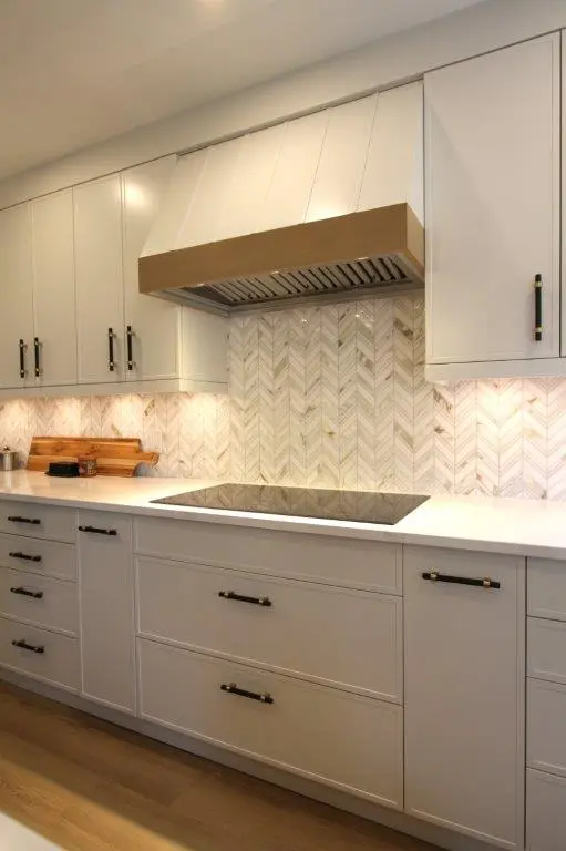 A modern kitchen with light grey cabinets, a white quartz countertop, and a herringbone tile backsplash. The kitchen features an induction cooktop and a stainless-steel range hood with a wood trim, with under-cabinet lighting illuminating the backsplash.