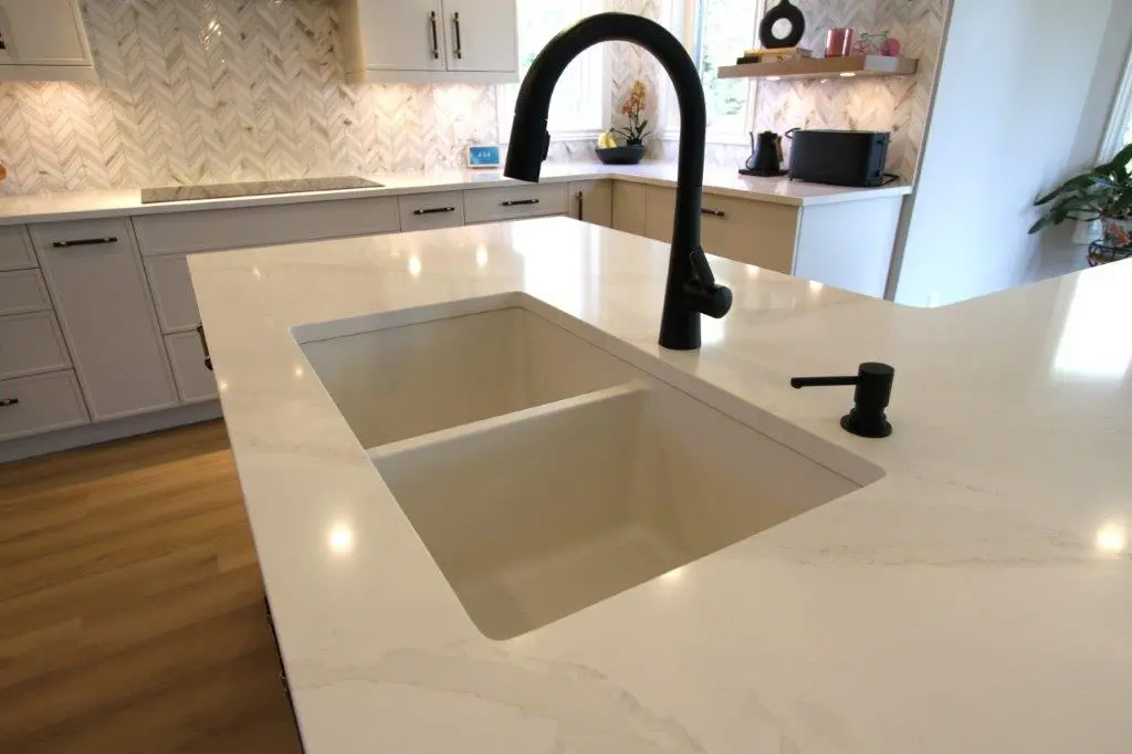 Modern kitchen with a white quartz countertop featuring a built-in sink with a black faucet, surrounded by light wooden cabinets and chevron-patterned backsplash tiles.