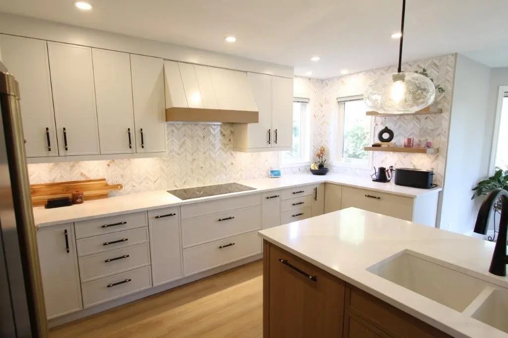 Modern kitchen with white cabinets, black handles, a large island with an inset sink, and chevron-patterned tile backsplash. Two pendant lights hang above the island, and there are two small windows on the wall near floating shelves.