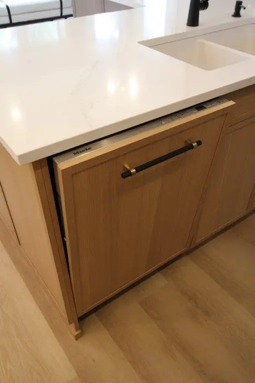 A kitchen island with a white countertop and a wooden panel dishwasher partially open, featuring a black handle and a sink with a black faucet nearby.