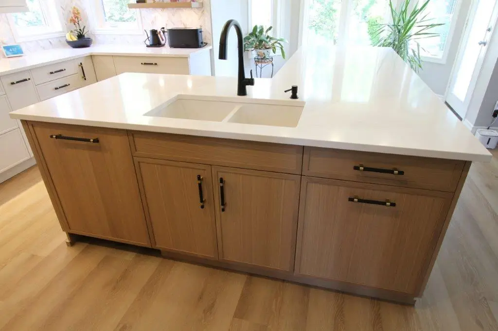 Modern kitchen island with a white quartz countertop, built-in sink, and brown wooden cabinetry, set in a bright kitchen with large windows and potted plants.