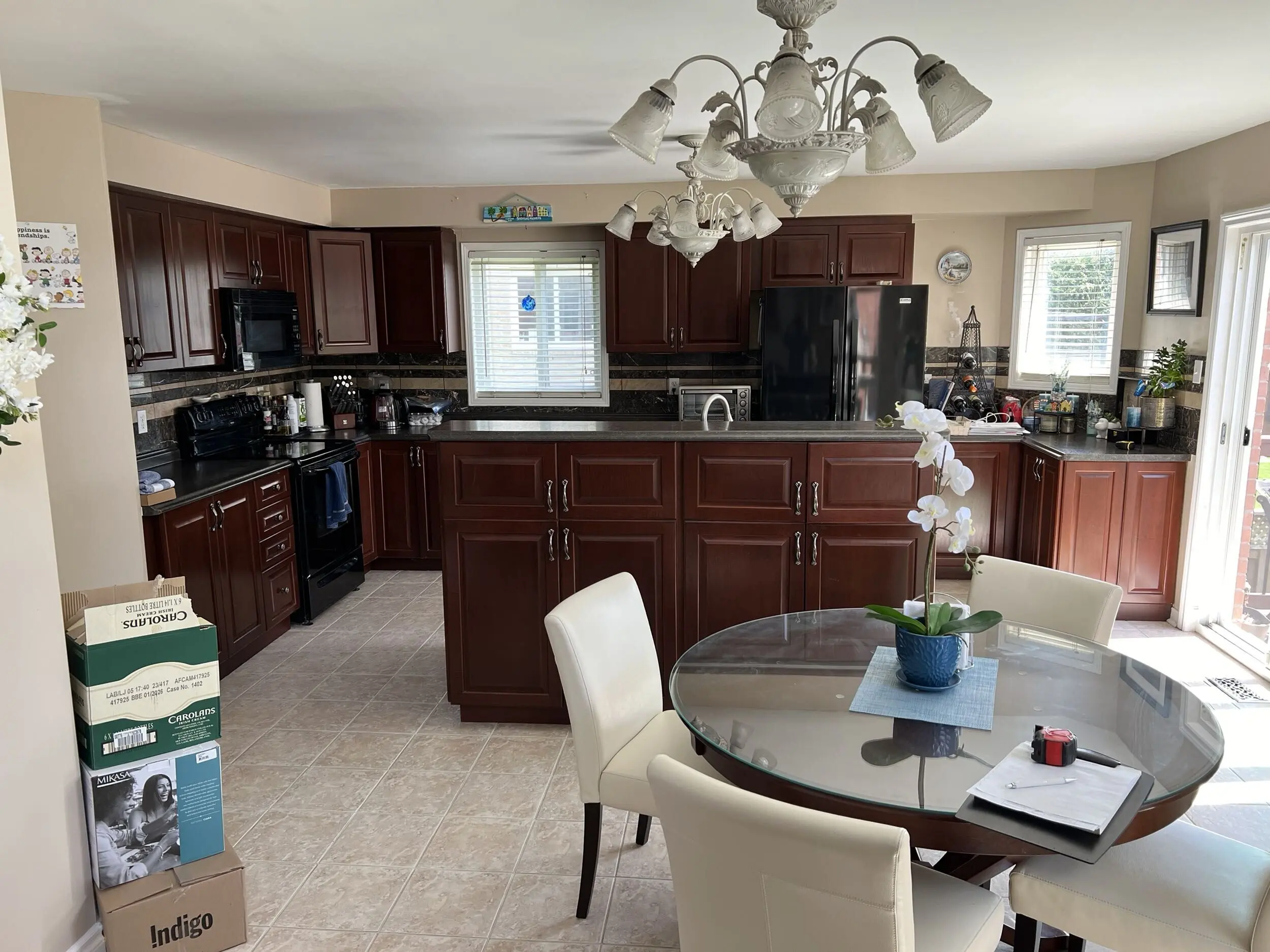 A before renovation kitchen with dark wooden cabinets, a black fridge, and a glass-top dining table with beige chairs. A plant in a blue pot sits on the table, which is set on a tiled floor. There is a window allowing natural light into the room.