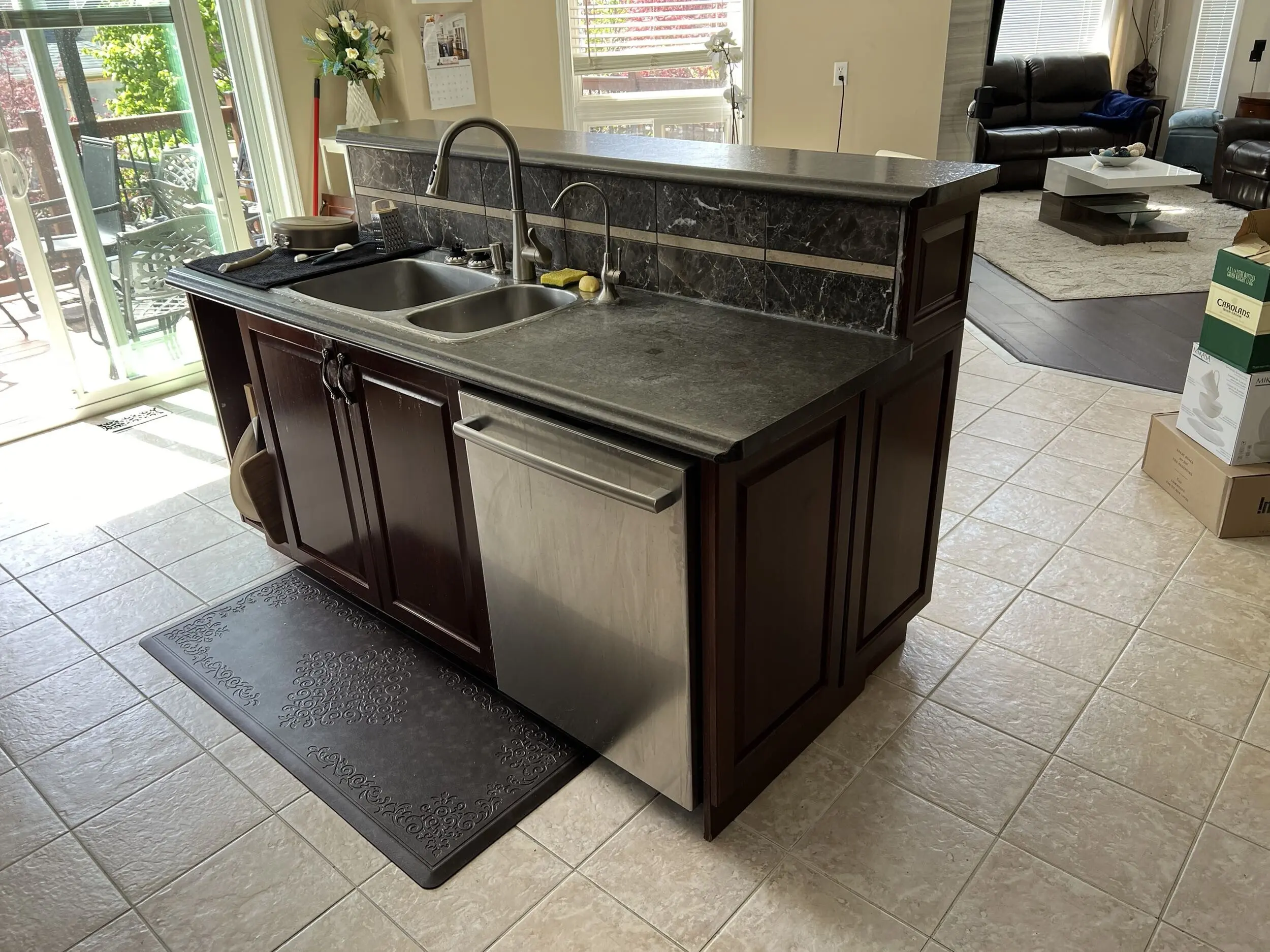 A before renovation kitchen island with dark wooden cabinets and a gray countertop featuring a double sink and modern faucet, located on a tiled floor, with a view of a living room and a patio through sliding glass doors.