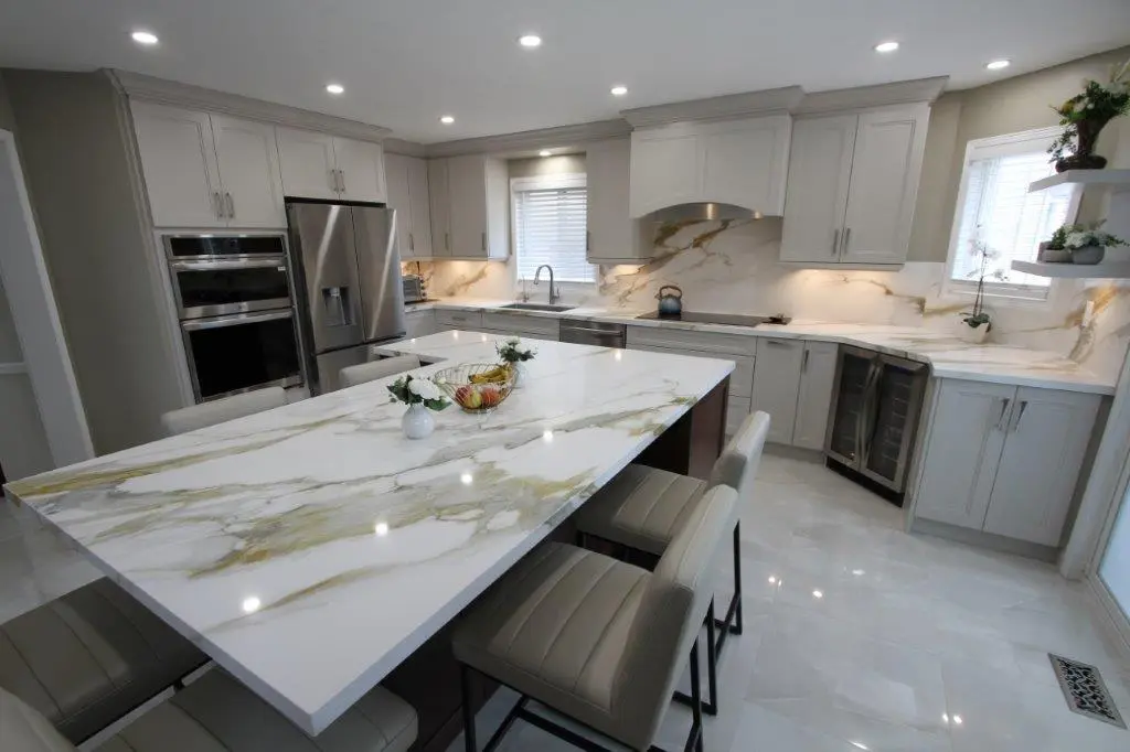 Modern kitchen with white and gray cabinetry, porcelain countertops, and a large island with seating. Stainless steel appliances, including a refrigerator and built-in ovens, are visible alongside a wine cooler. The room is well-lit with recessed lighting and decorated with potted plants.