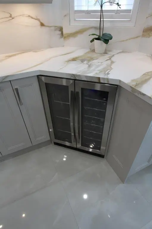 A modern kitchen corner featuring a small wine fridge with double glass doors under a porcelain countertop, accompanied by a potted plant in front of a window.