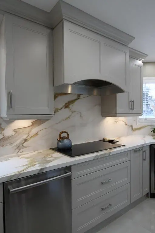 Modern kitchen with light gray cabinets, a porcelain backsplash, and a black electric cooktop. A blue kettle is placed on the cooktop, with natural light coming through a window on the right.